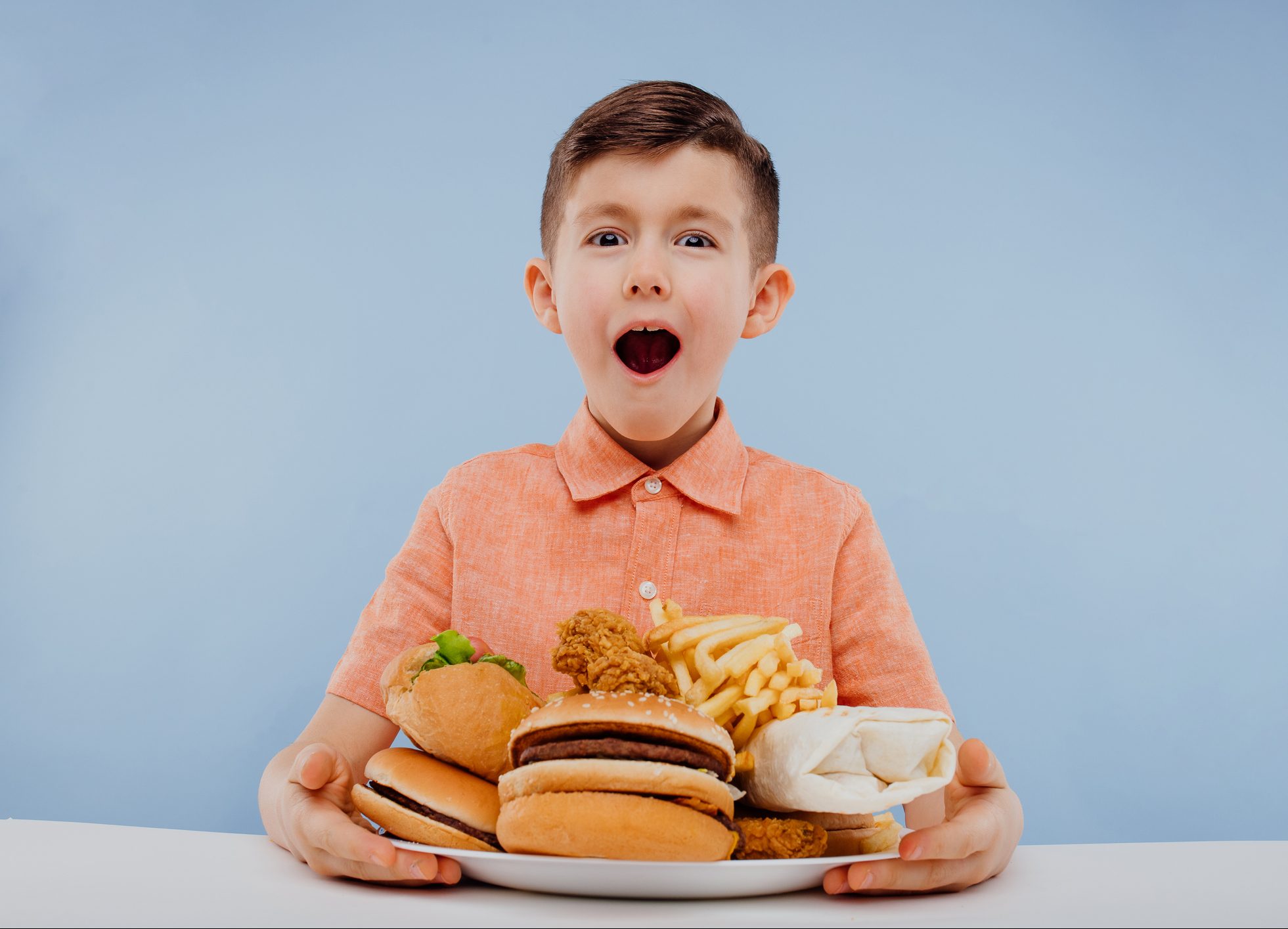 FUNIBER-excited-little-boy-with-open-mouth-has-junk-food-on-the-table-looking-forward-isolated-on-blue-background-studio-